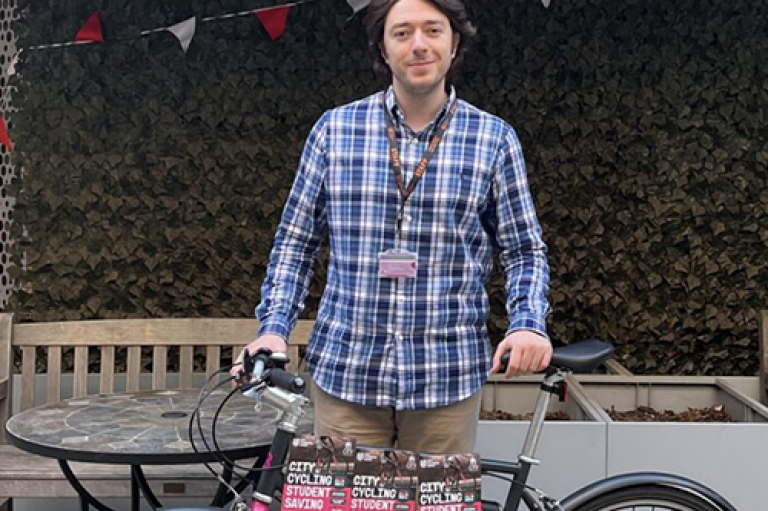Yavuz Kafadar, Deputy President of City Student's Union, stands outside with a bike. He is wearing a blue shirt and beige trousers. There is colourful bunting in the background.