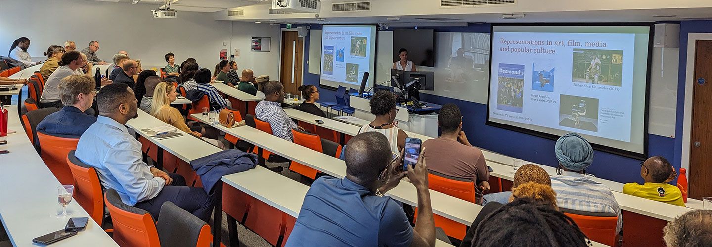 A group of people sit at rows of desks in front of a screen with a slide which reads 'Representations in art, film, media and popular culture'. There are image screenshots of film and books. 