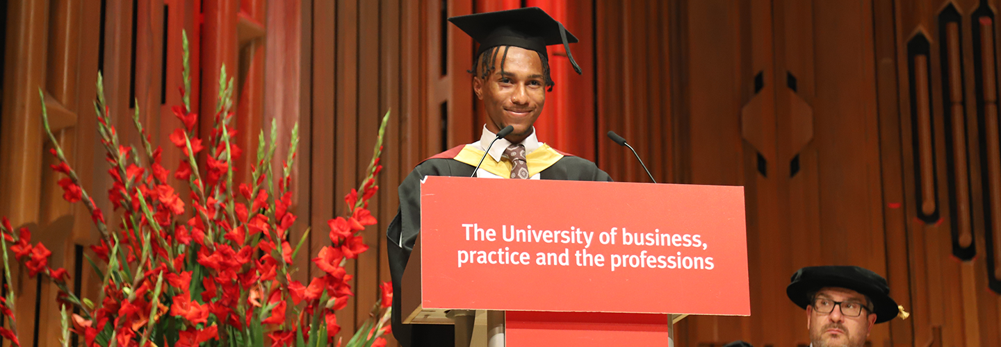 A person wearing a graduation cap and robe smiles in front of a microphone and podium