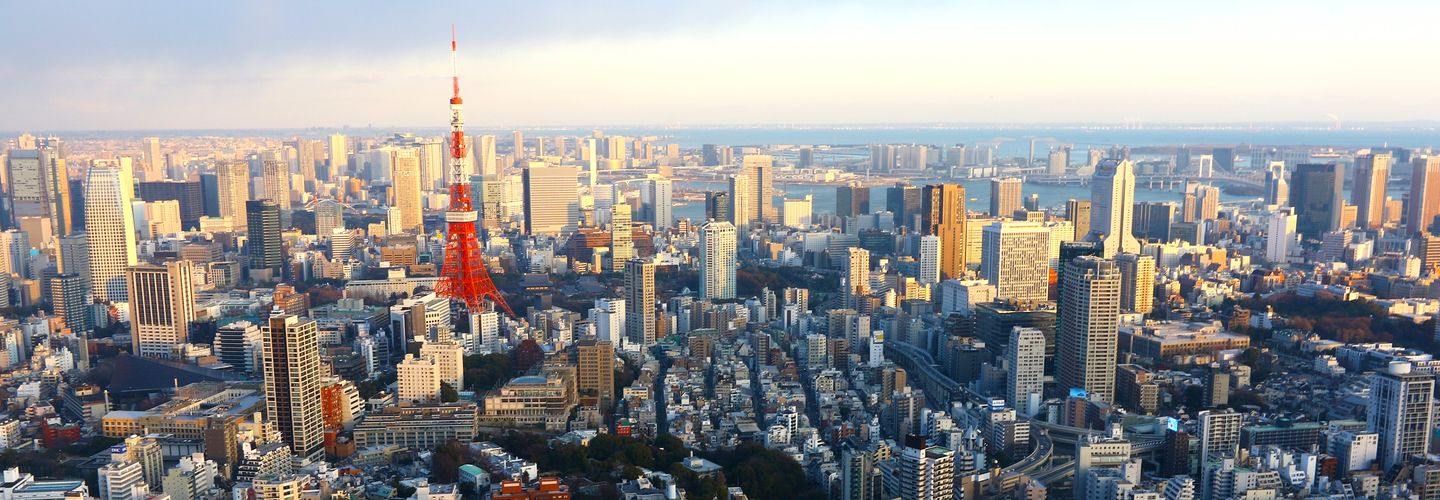 An image of the skyline of Japan. In the distance is the sea, to the forefront, the red and white Tokyo Tower rises above the highrises around it.