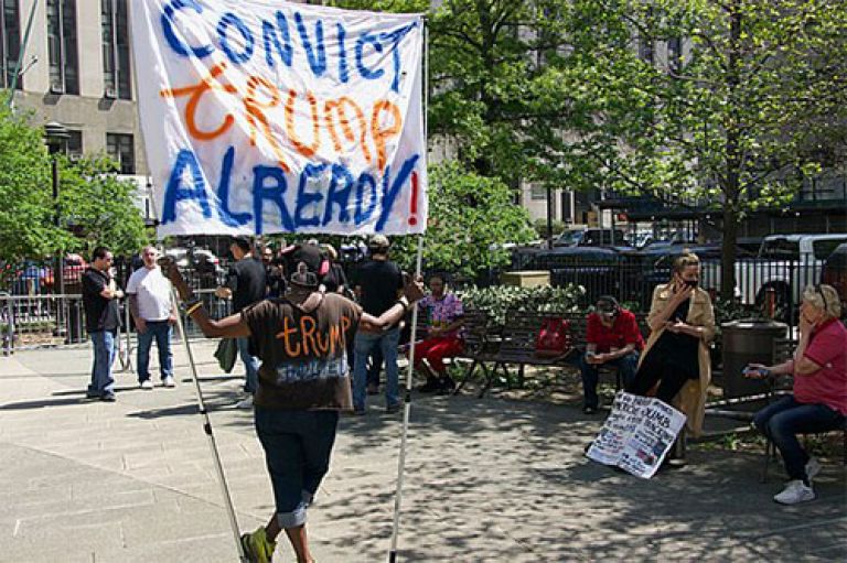 Image of a protestor outside of Trump's court trial for paying hush money to pornstar Stormy Daniels. The Protestor holds a banner which reads 'Convict Trump Already!'. Around him, other protesters sit on benches and mill in a sunny square with trees in the background
