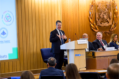 A man stands at a lectern with the Bayes logo with a picture of men in 1970s fashion in the background