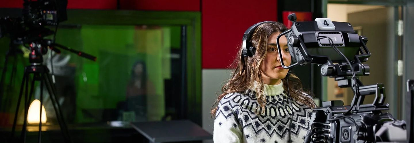 An image of a student with shoulder length brown hair and white jumper looking into the lens of a large camera. In the background is a second, blurry camera