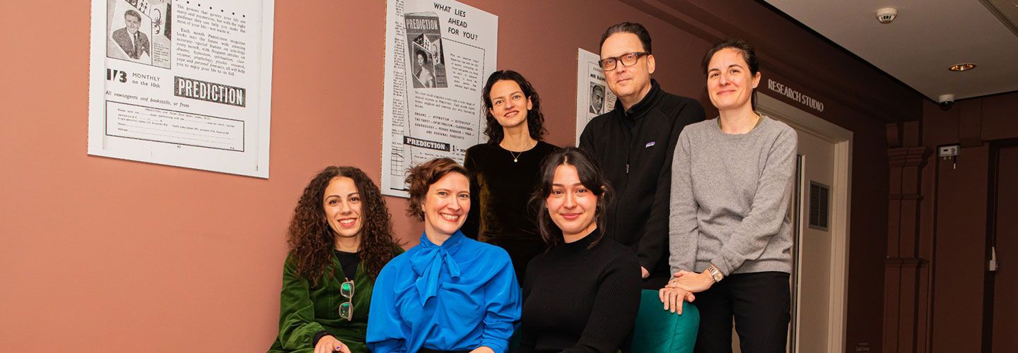 An image of Dr Lise Butler and her co-investigators sitting on a green armchair and others standing at the Manchester Museum. They are in a room that has walls that are a light pink / salmon colour, and a lighter similar colour on the ground. Behind them are two white posters against the wall that look like newspapers with pictures. We can read the words 