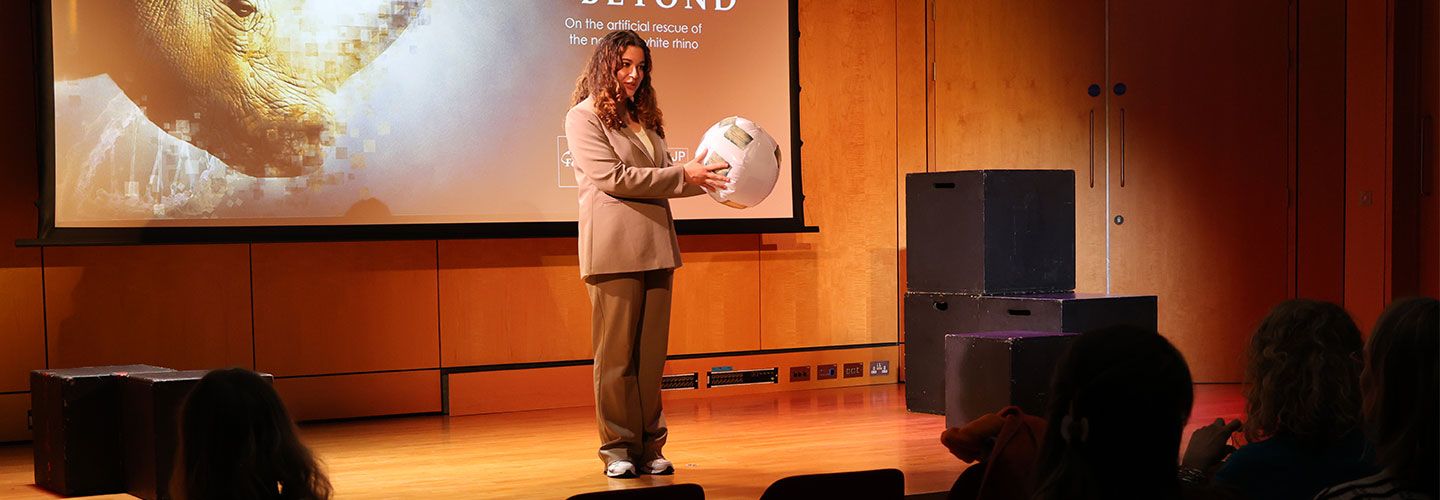 Journalist Jasmijn Apte wears a beige suit and stands on stage. She holds a beach ball. Behind her can be seen a screen which projects an image of a rhino and the title of her performance 