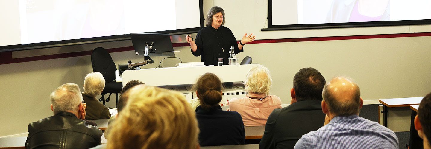 An image of Prof Wendy Pullan standing at with her hands out to either side. In front of Wendy, an audience sits in rows in an amphitheatre-style lecture room.