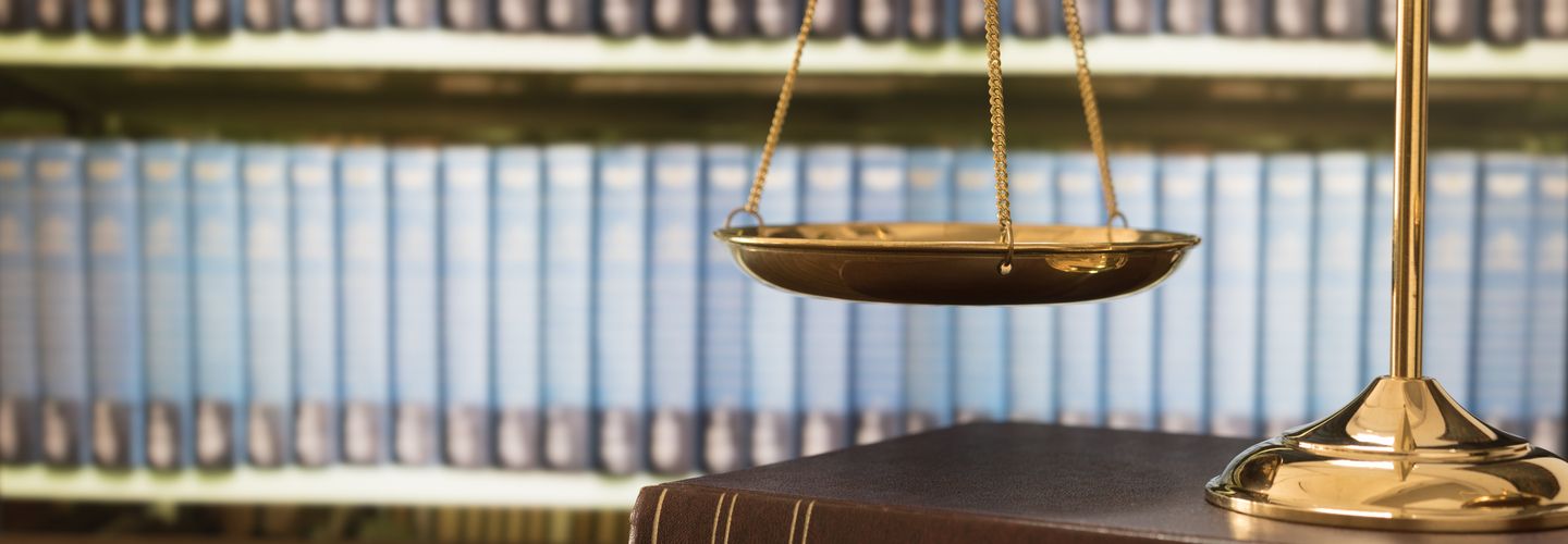 An image of golden scales, representing scales of justice, on top of a dark brown book. In the background, blurry, are rows of books in a library of a law firm