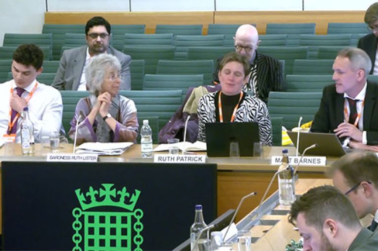 Experts sit in a row on a light brown wooden table with laptops, water, notebooks, microphones and their names. Behind them, people sit in green chairs to listen. The experts are sitting in the Boothroyd room in the House of Commons