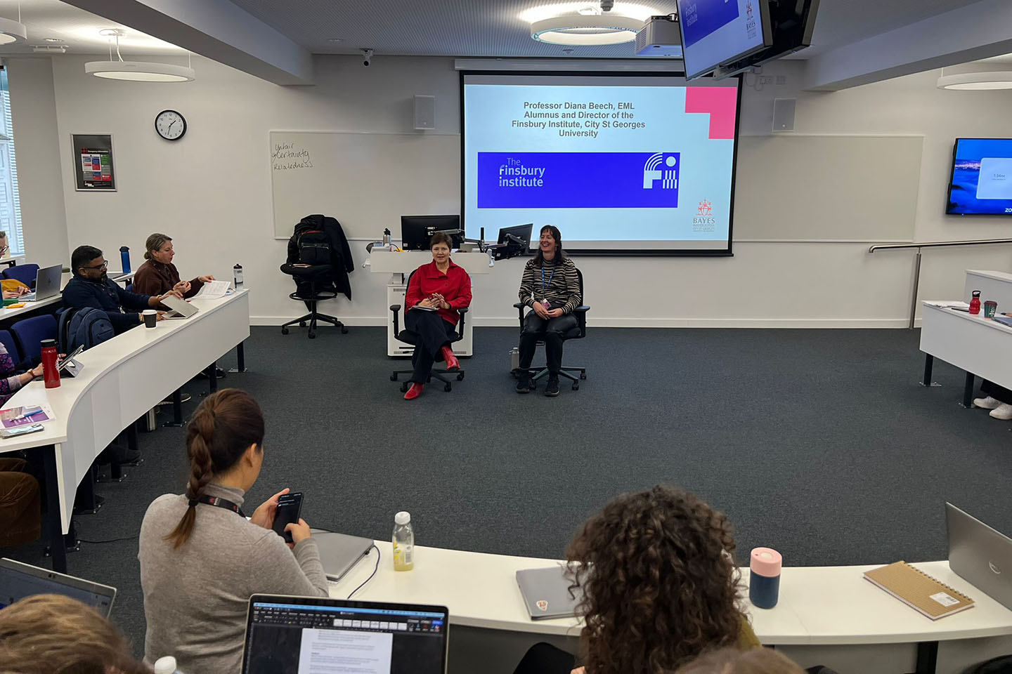Two women sit in front of a presentation screen, with an audience in a lecture theatre.