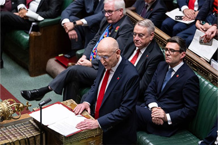 An image of Chancellor Healey in the House of Parliament delivering the Budget. He wears a smart dark blue suit and red tie and small poppy pin on his suit. Sitting behind him can be seen PM Burnham. In front of him is the corner of a crown