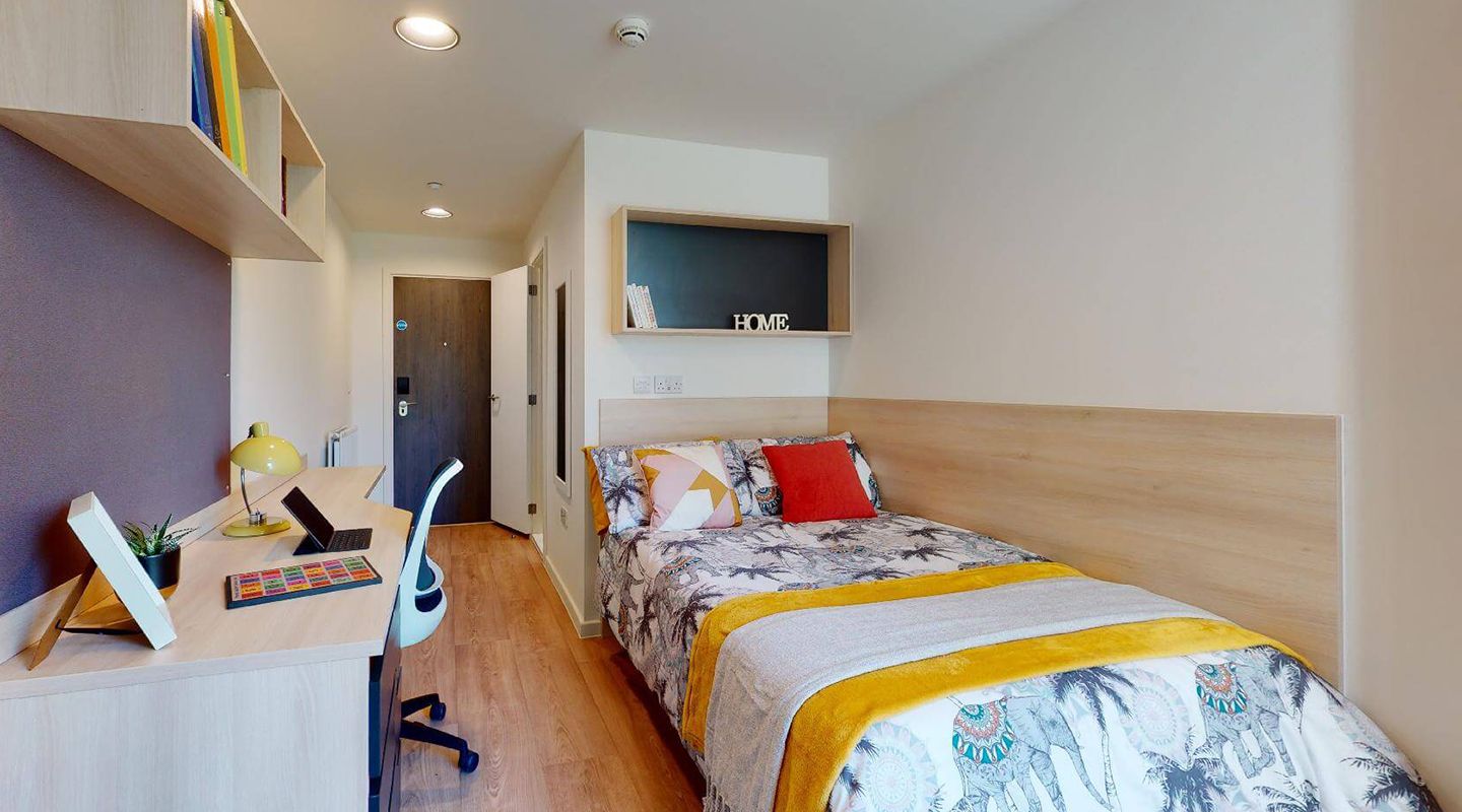 Single bed and desk. Pale laminate floor, cream walls and furniture in pale wood and black. Radiator and mirror by the door at the far end. En-suite bathroom behind the bed, unseen but with door open.