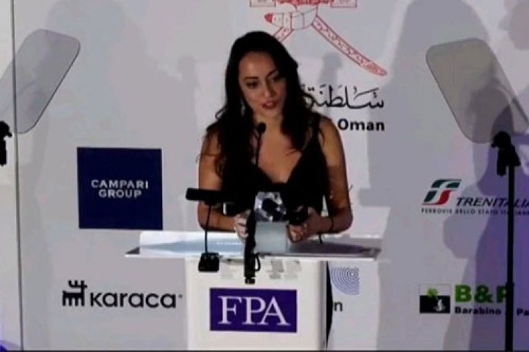 Maya Saad, MA Investigative Journalism alumna (2023), stands in front of a lectern with the letters FPA on it as she receives an award at the Foreign Press Association 2023 awards. She wears a smart black dress and has her long hair down. Behind her 