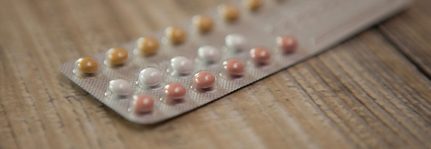 Image of a pack of contraceptive pills (a row of yellow, then a row of white, then a row of pink pills) against a wooden table
