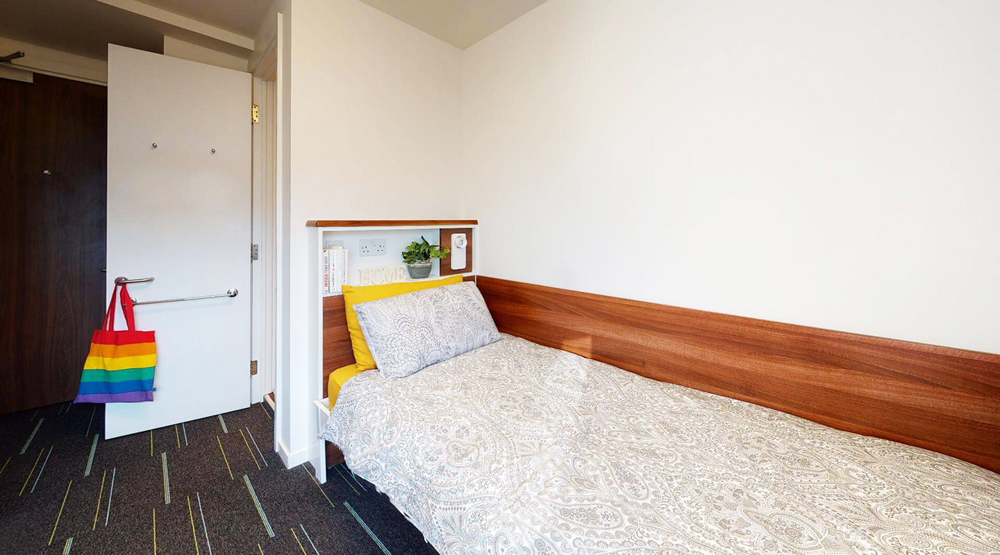 Bedroom with a single bed against the wall and a built in headboard. The bathroom door is seen open immediately beyond the bed, and past that is the front door.