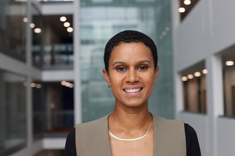 A headshot of Dr Karis Campion smiling to camera. She has a shaved head and wear a smart chain and dress. The background is blurry behind her