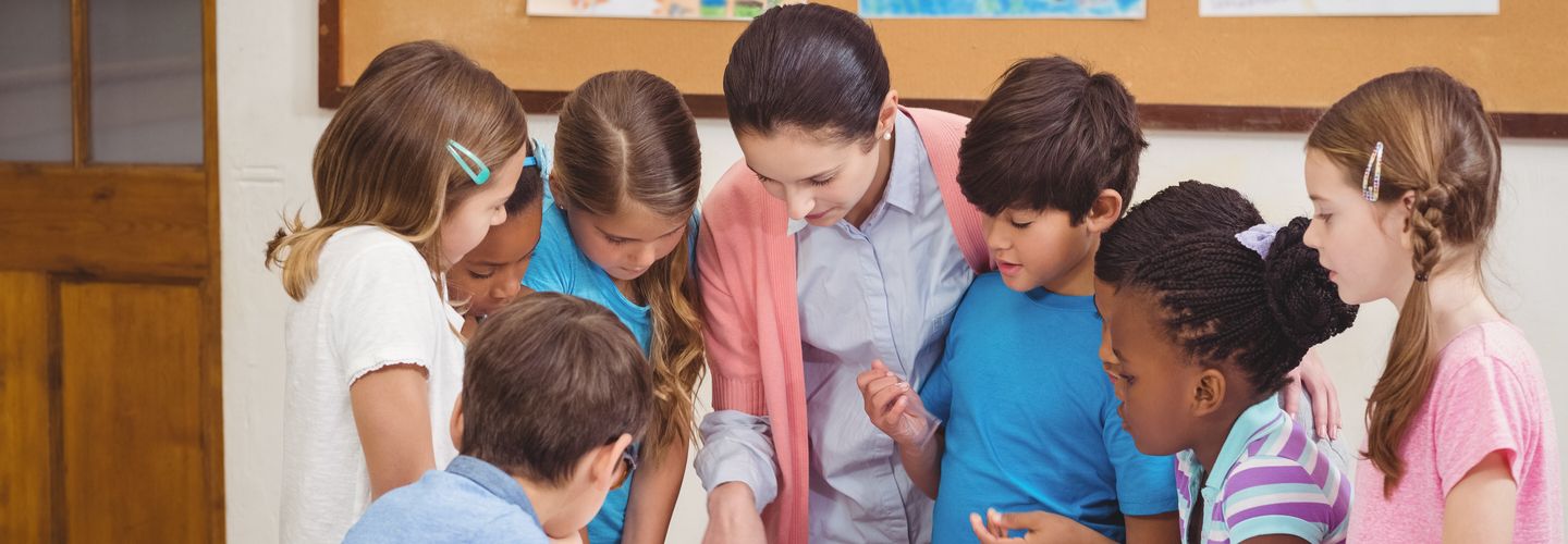 A teacher stands among a diverse group of schoolchildren at a desk and points to an open notebook. Behind them, you can see a blurry background with children's drawings