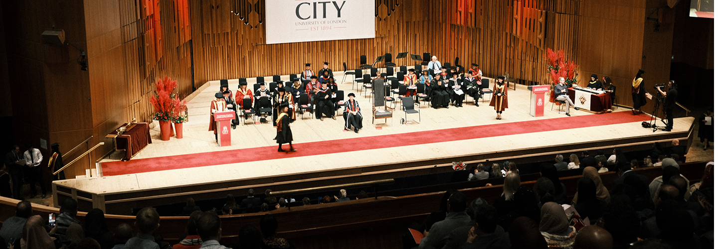A distant shot of a person at a lectern with a red background featuring the City logo