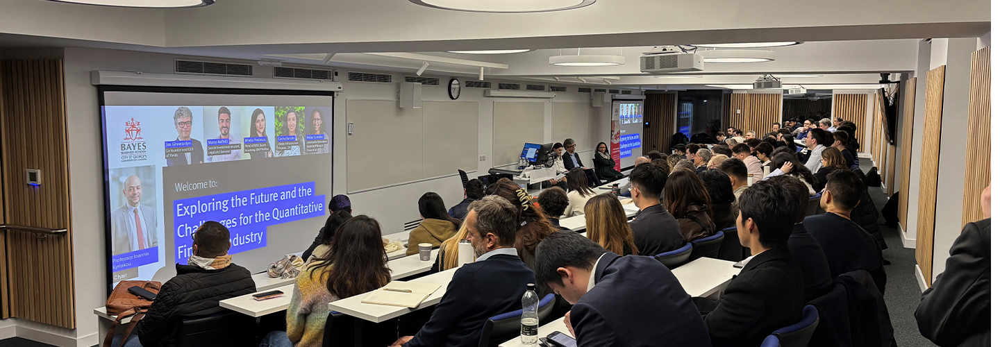 A lecture theatre with dozens of people and a screen showing the speakers in a panel discussion to start soon