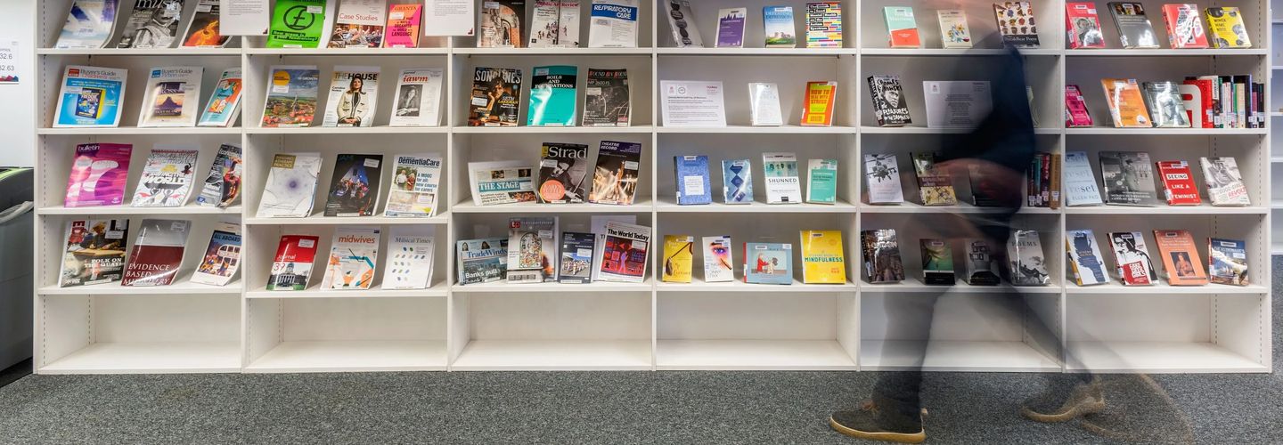 An image of a bookshelf with books on display. A blurry figure walks in front of it