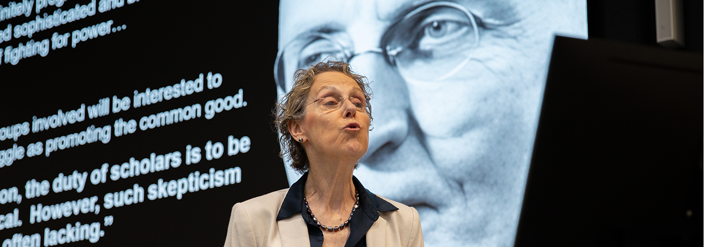  A woman on a stage pursing her lips while making a point while behind her on a screen there is a black and white picture of an elderly man with a quote out to the side