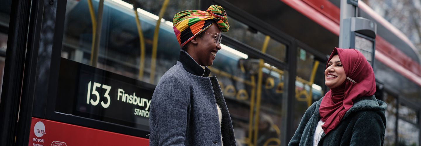 Two young women talking cheerfully as the number 153 bus pulls up behind them.
