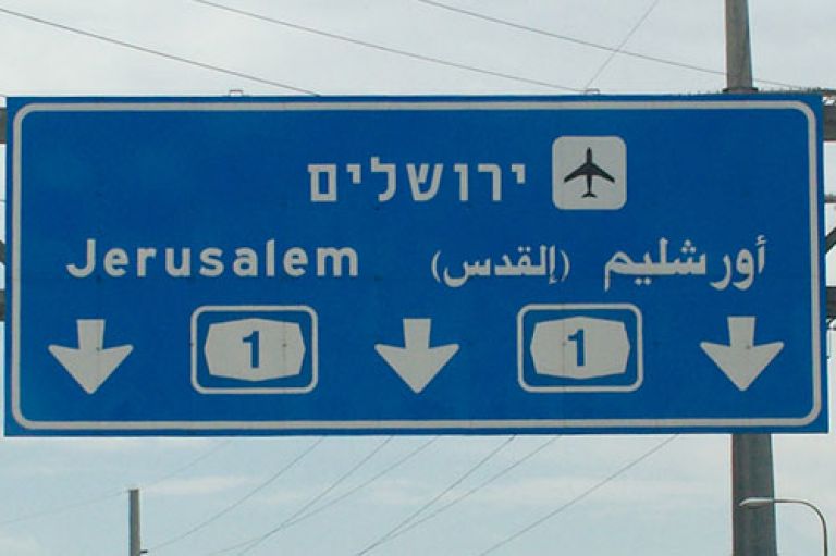 An image of two signs on the motorway in Israel, one is blue and points towards Jerusalem and the airport. The other is green and yellow and points towards a Kibbutz. Some of it is written in Hebrew. 