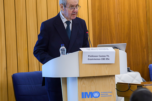 A man stands at a lectern with the Bayes logo with a picture of men in 1970s fashion in the background