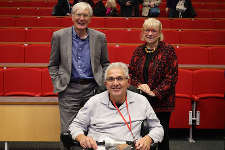 David Walker and Polly Toynbee stand at smile to camera in front of rows of red seats. In front of them sits Prof Petros Iosifidis in his wheelchair, who is holding a microphone in his lap