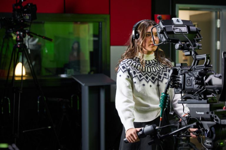 An image of a student with shoulder length brown hair and white jumper looking into the lens of a large camera. In the background is a second, blurry camera