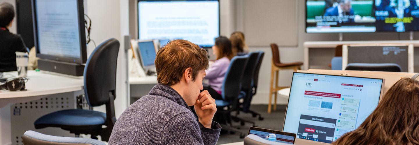 Students working in computer room with large screen displaying news overhead.