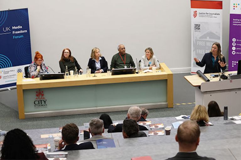 Five speakers sit in a row at the front of an amphitheatre style lecture room. In front of them are glasses of water and small microphones. To their right, standing at a standing desk is Prof Mel Bunce, gesturing towards the audience. Behind them are three large banners, one for the Media Freedom Forum, one for the Foreign Policy Centre and one for the Centre for Justice for Journalists