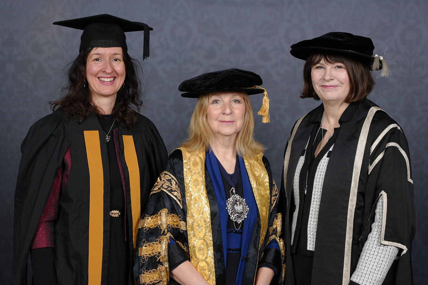 Three women pose together in academic robes and caps. The woman in the centre wears a black gown embroidered and edged with gold, and a large filigree medallion.
