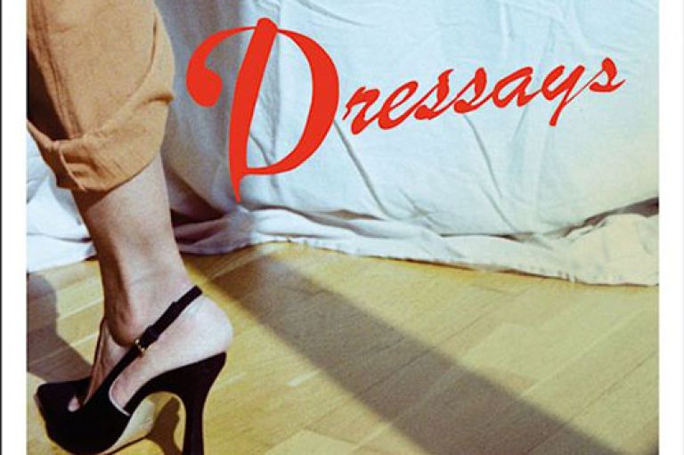 An image of the cover of Dressays. We can see a woman's legs, wearing camel trousers and putting on a pair of black slingback heels. He stands on a floor with wooden slats, and behind her is a sheet covering a wall. To the top right are the words 