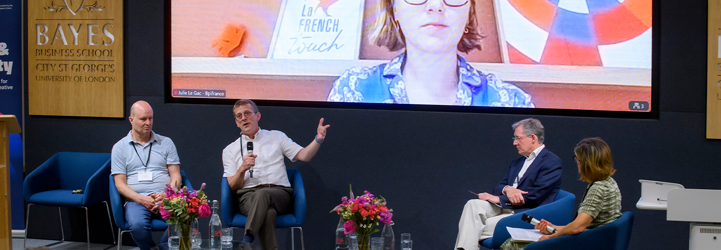 Three men and a woman on a stage with flowers with one man using a microphone and a woman on a screen in the background