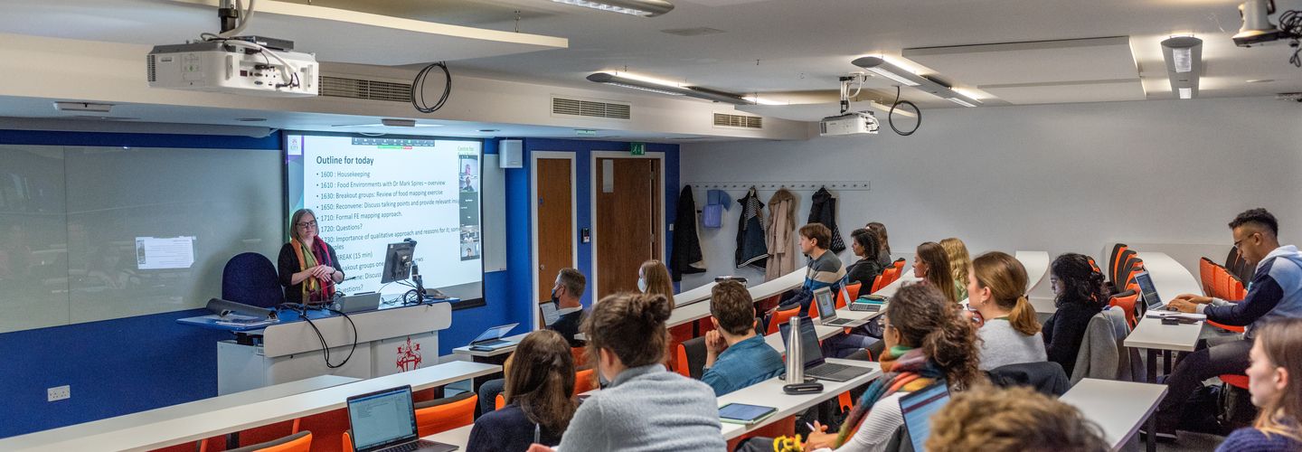 A lecturer stands at a podium next to her presentation display, speaking to students in the lecture theatre.