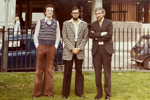 Three men in 1970s clothes including flared trousers and a tank top and with big sideburns outside a building