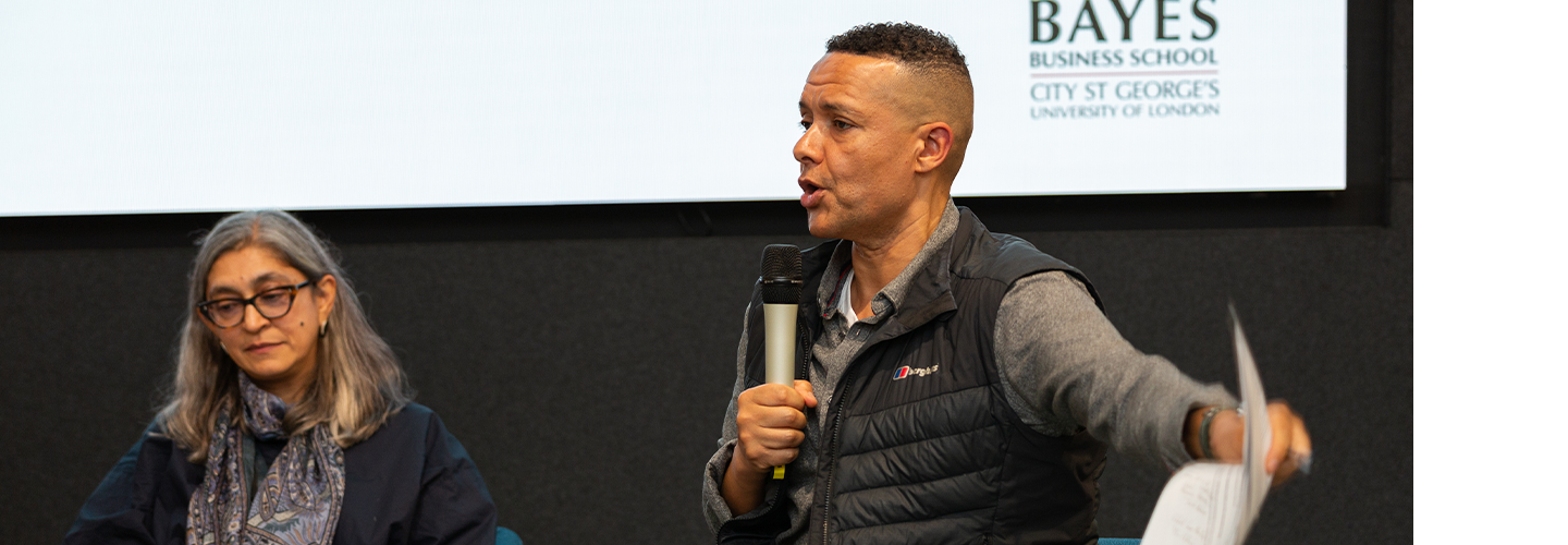 A man with a microphone talks while a woman listens in front of a board with Bayes Business School on it