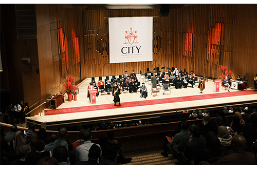 The City logo against a red backdrop on a stage with a person at some distance speaking at a lectern