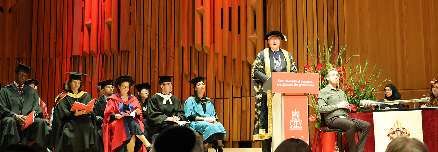 A man stands on stage at a podium to address an audience, with people dressed in academic bonnets and robes joining him on stage
