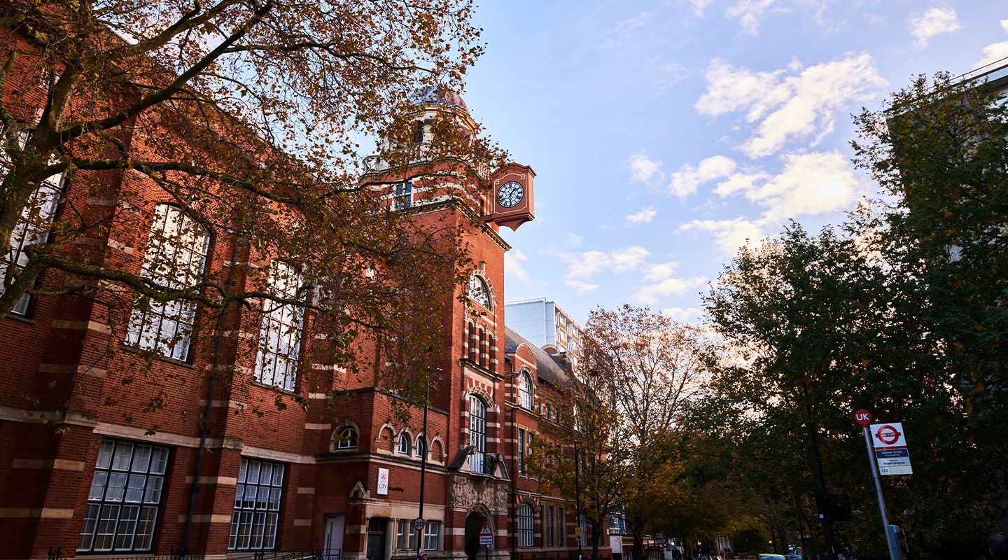 City's red-brick college building with a clock tower, surrounded by trees with autumn leaves.