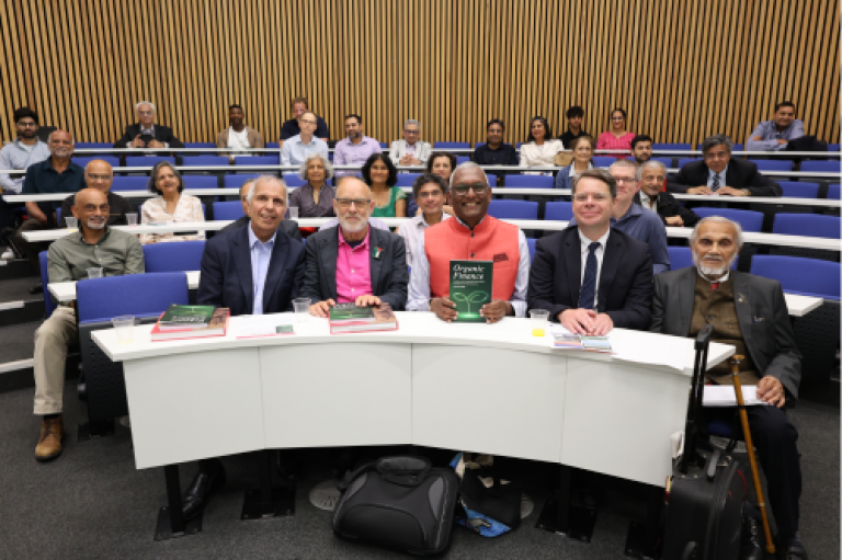 The five panellists sit at a rounded desk and smile to the camera. In front of them are copies of the book 