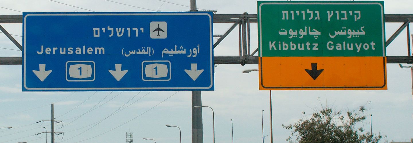 An image of two signs on the motorway in Israel, one is blue and points towards Jerusalem and the airport. The other is green and yellow and points towards a Kibbutz. Some of it is written in Hebrew.