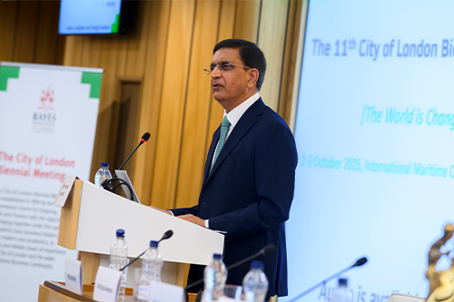 A man stands at a lectern with the Bayes logo with a picture of men in 1970s fashion in the background