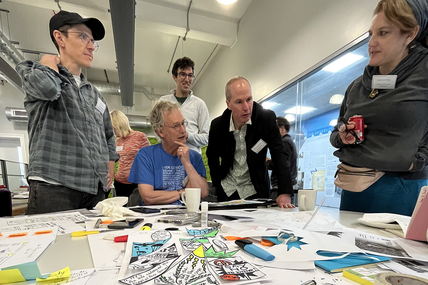 Group in discussion gathered around a table cluttered with paper, pens and drawn comics.