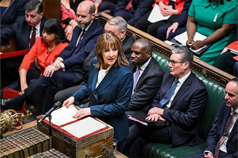 An image from above of Chancellor Reeves standing at a lectern and reading her Autumn Budget statement in the House of Commons. Behind her sit Prime Minister Keir Starmer and front benchers.