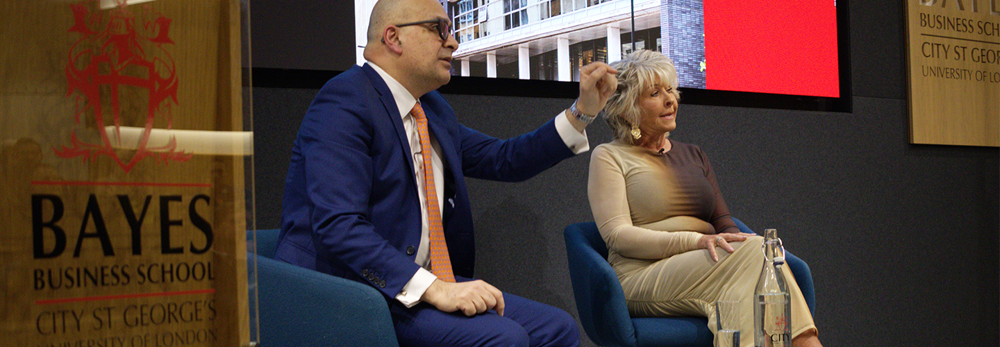 An Asian man and a white woman sit in chairs looking out at the audience with Bayes Business School branding around them