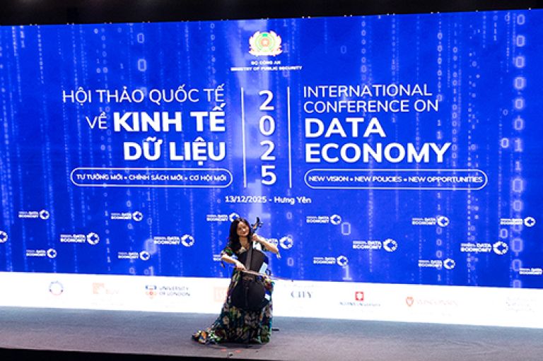A woman sits playing a cello in front of a brightly lit screen