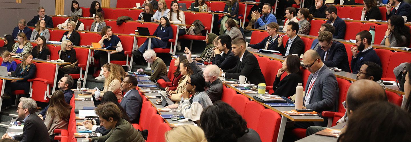 An audience member asks a question. He sits around a full audience sitting on rows of red chairs. 