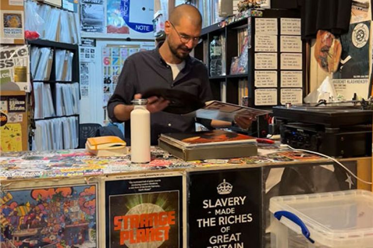 Dr Lmabros looks down at a turnstile and smiles. He is in a record shop with lots of posters and stickers of various musicians and with political slogans all around him.
