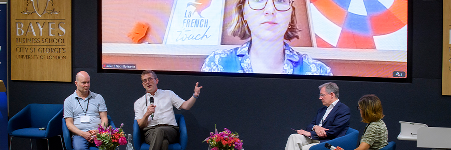 Three men and a woman on a stage with flowers with one man using a microphone and a woman on a screen in the background